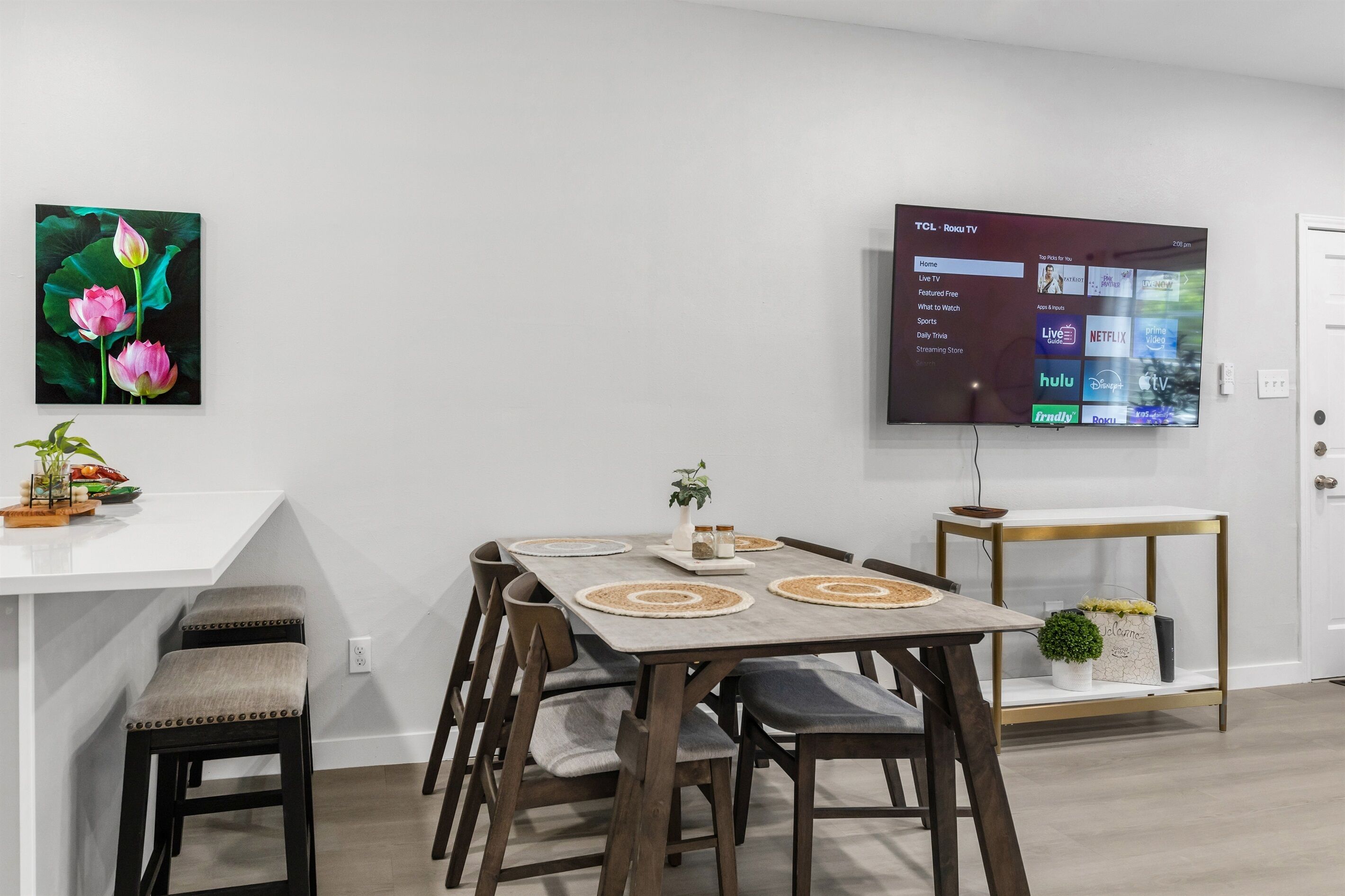 A modern dining area with a white table, wooden chairs, and a flat-screen TV mounted on the wall displaying a streaming service interface.