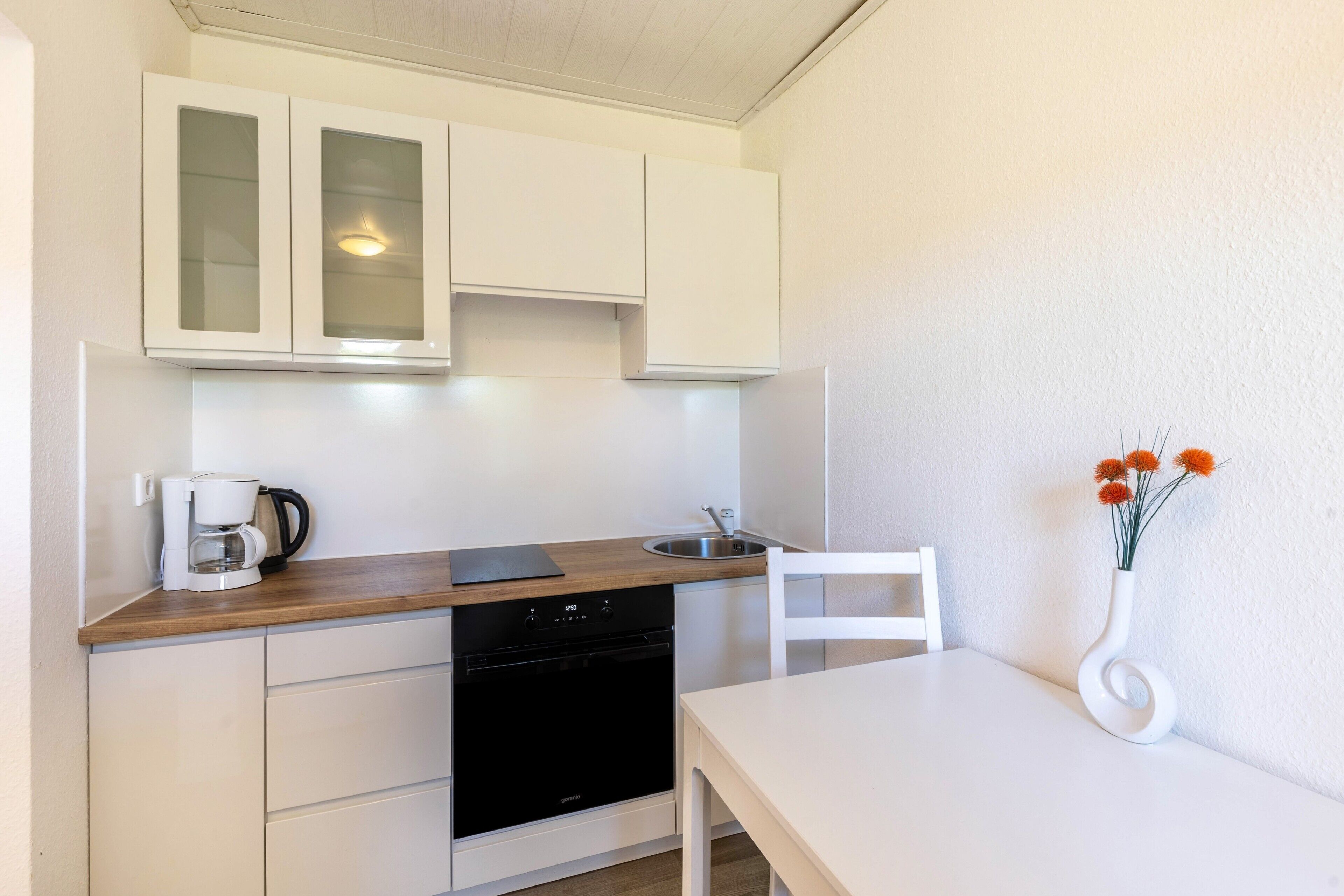 Une cuisine avec des meubles blancs, un lave-vaisselle, une machine à café, une chaise blanche et un vase rempli de fleurs.