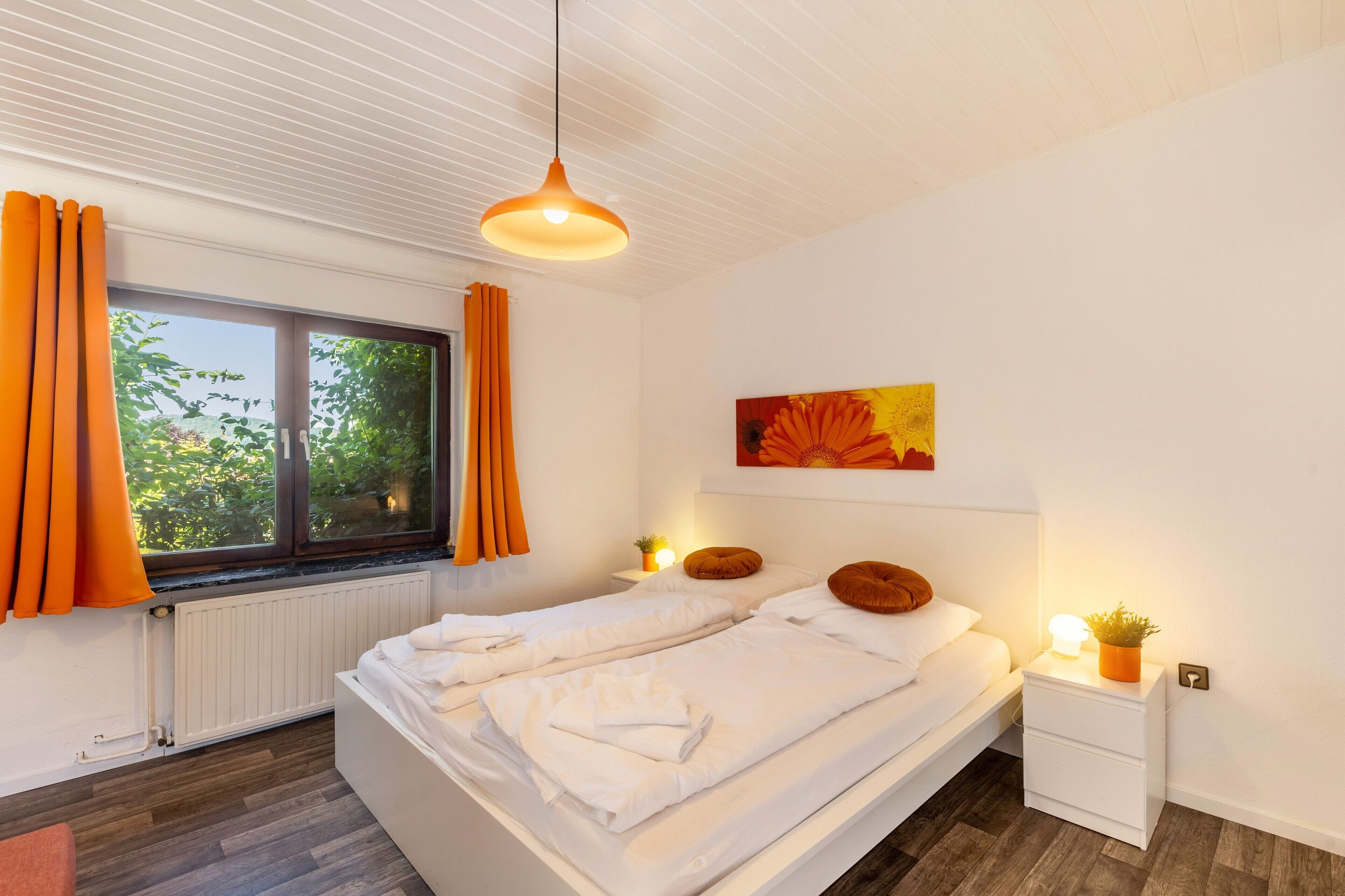 A bedroom with two beds, a window with curtains, a radiator, a bedside table with a lamp, and a painting on the wall.