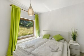 Ein Schlafzimmer mit Bett, grünen Vorhängen, einer Zimmerpflanze und einem Fenster mit Blick auf Grünfläche.