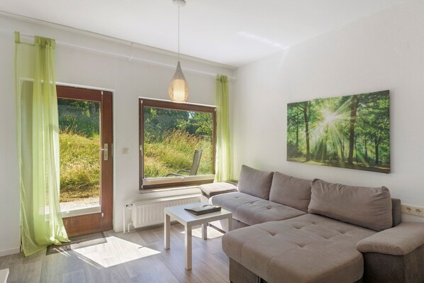Un salon moderne avec un canapé, une table, une suspension et une grande fenêtre donnant sur un paysage verdoyant.