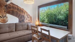 A room with a large giraffe mural, a sofa, a dining table, chairs, a hanging light fixture, and a window with a view of trees. - Holiday Home 'Giraffe' with Private Terrace and Wi-Fi (Niedenstein)