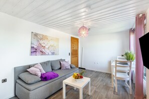 A modern living room with a grey sofa, a white coffee table with fruit, a wooden door, and a pink pendant light. - Holiday Home 'Die Aussicht' with Mountain View, Private Terrace & Wi-Fi (Niedenstein)