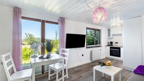 A modern living room with a dining table, white chairs, a flat-screen TV, and a kitchen area with a microwave and oven.