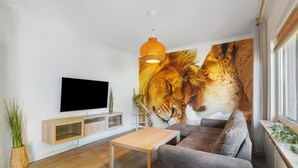 A living room with a flat-screen TV, a sofa, a coffee table, a wall-mounted TV stand, and a large lion mural. - Holiday Home 'Löwe' with Private Terrace and Wi-Fi (Niedenstein)