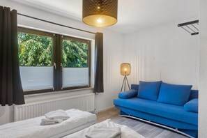 A modern room with a bed, a blue sofa, a standing lamp, and a window with curtains. - Bungalow 'Leopard' with Shared Terrace, Shared Garden and Wi-Fi (Niedenstein)