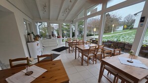 Un restaurant avec des tables et des chaises en bois, un espace cuisine et de grandes fenêtres donnant sur l’extérieur.