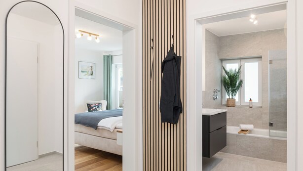 A modern bedroom with a bed, a bedside table, and a nightstand. There is a bathroom with a bathtub, a sink, and a mirror. The room has a wooden floor and a white wall with a vertical wooden slat design. - FENJOY vacation apartment No°9 with terrace | garden | BBQ (Bernau am Chiemsee)