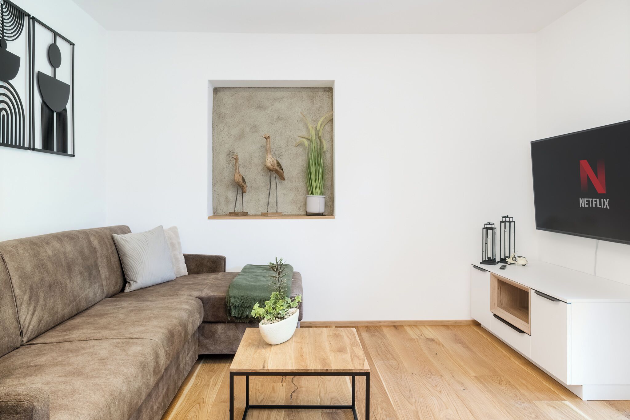 A modern living room with a brown sofa, a coffee table with a plant, a wall-mounted artwork, and a white entertainment unit with a Netflix logo.
