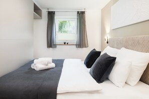 Cama bem arrumada com travesseiros brancos e escuros, uma toalha dobrada e uma janela com cortinas.