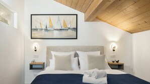 Een slaapkamer met een bed, hoofdbord, nachtkastjes, wandlampen en een lijst met een afbeelding van zeilboten.