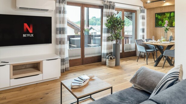 Een moderne woonkamer met een flatscreen televisie, een bank, een eettafel met stoelen en een groot raam met gordijnen.