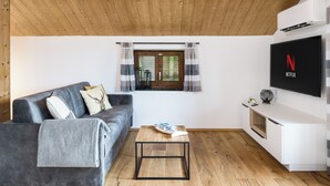 Een moderne woonkamer met een grijze bank, een houten salontafel, een aan de muur bevestigde televisie en een raam met gordijnen.