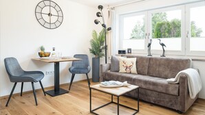 Een moderne woonkamer met een houten vloer, een grijze bank, twee fauteuils, een salontafel, een wandklok, een kamerplant en een raam met uitzicht op bomen.
