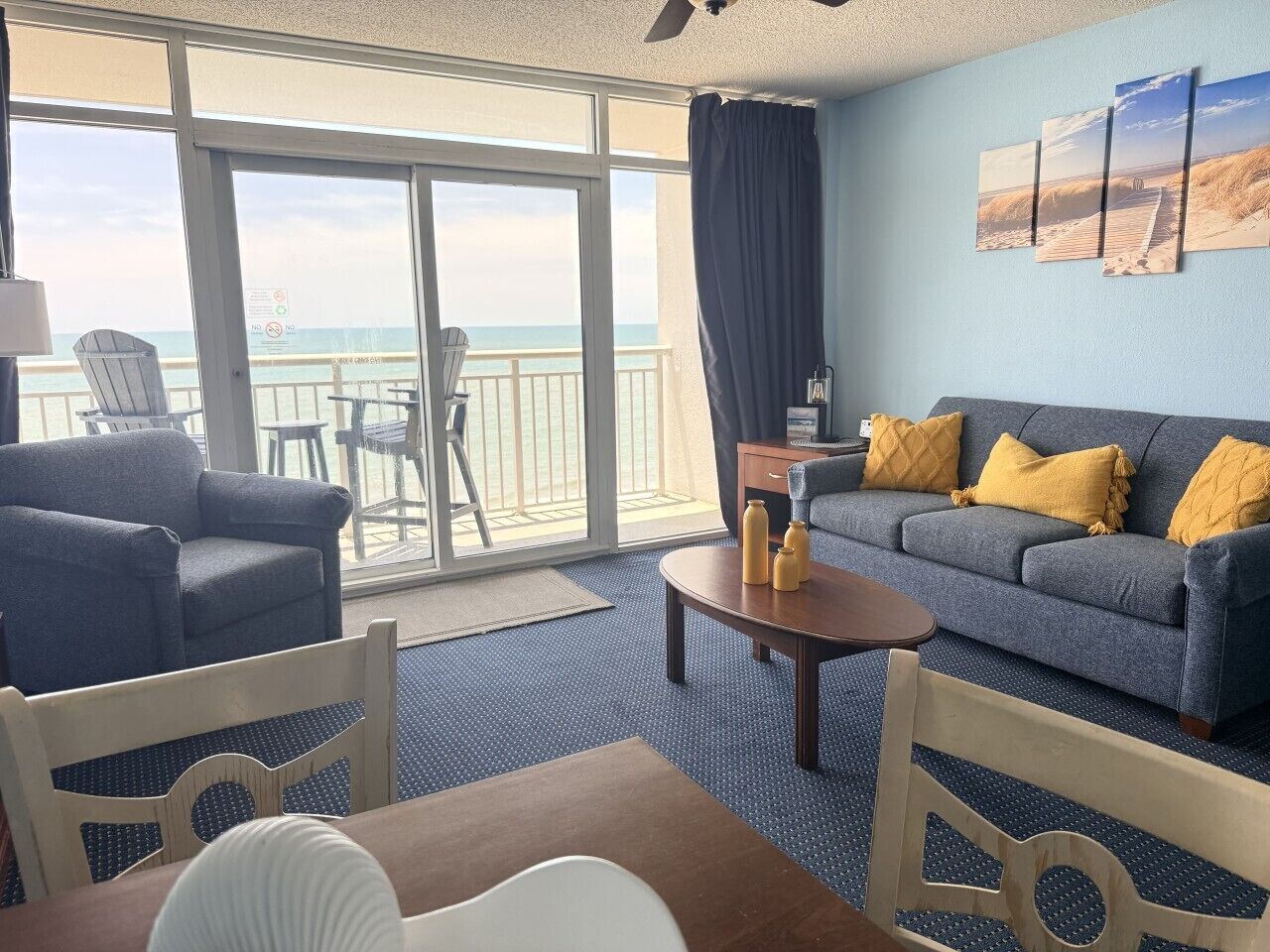 A room with a sofa, chairs, a table, and a view of the ocean through large windows.