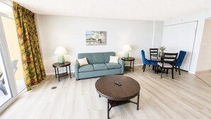 Una sala de estar con sofá, mesa de centro y sillas, un cuadro en la pared y una ventana con cortinas.