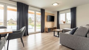 A modern living room with a sofa, a dining table with chairs, a flat-screen TV mounted on a wooden wall, and large windows with curtains.