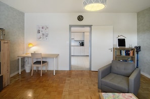 A room with a wooden desk, a chair, a sofa, a lamp, a bookshelf, and a door leading to a kitchen.