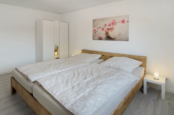 A bedroom with a wooden bed, white bedding, a bedside table with a lamp, and a wall-mounted artwork.