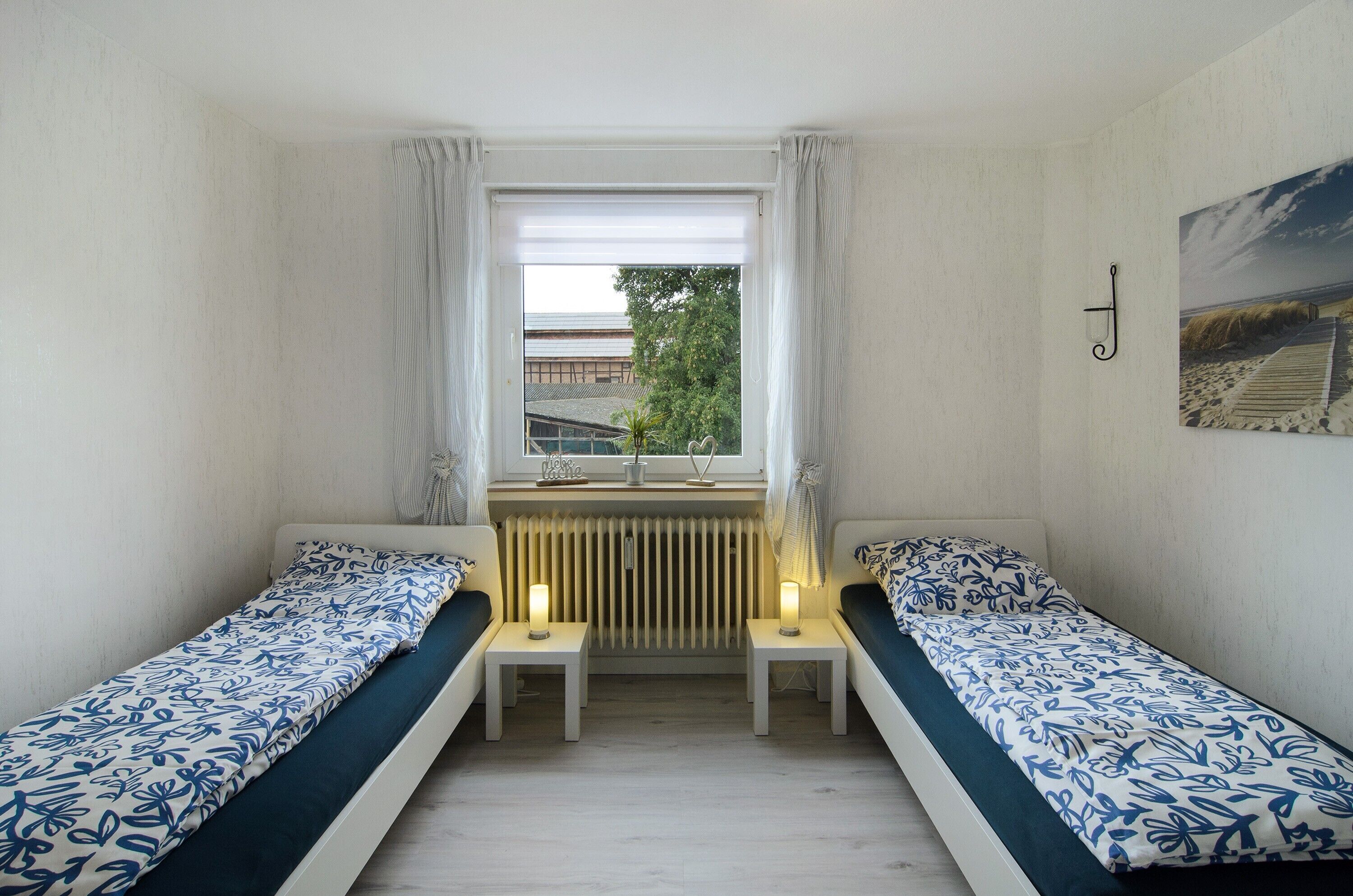 Una habitación pequeña con dos camas individuales, un radiador, una ventana con cortinas y vista a un edificio.