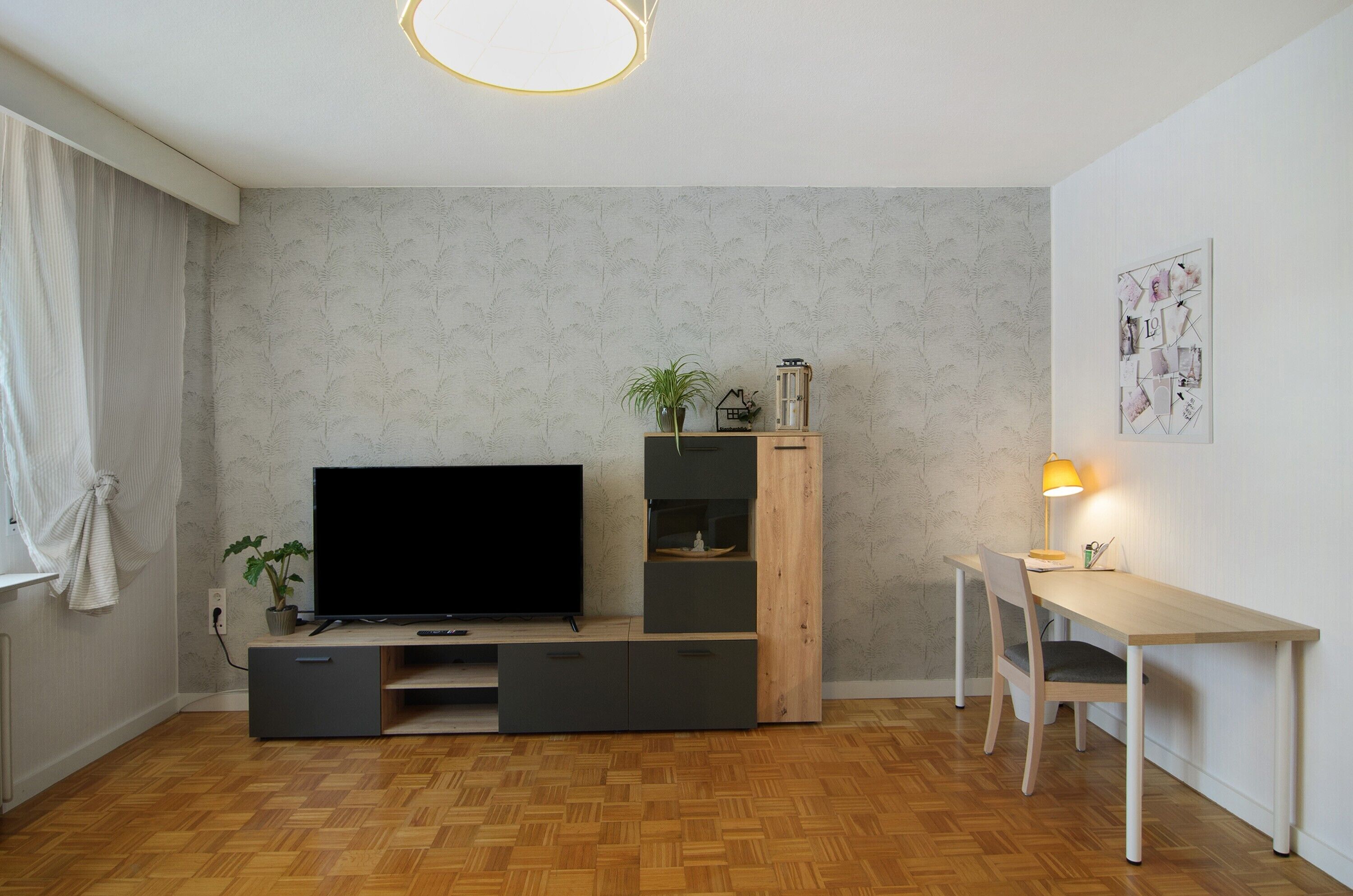Un salon moderne doté d’un téléviseur à écran plat, d’un meuble de rangement en bois, d’un bureau avec une chaise et d’une lampe fixée au mur.