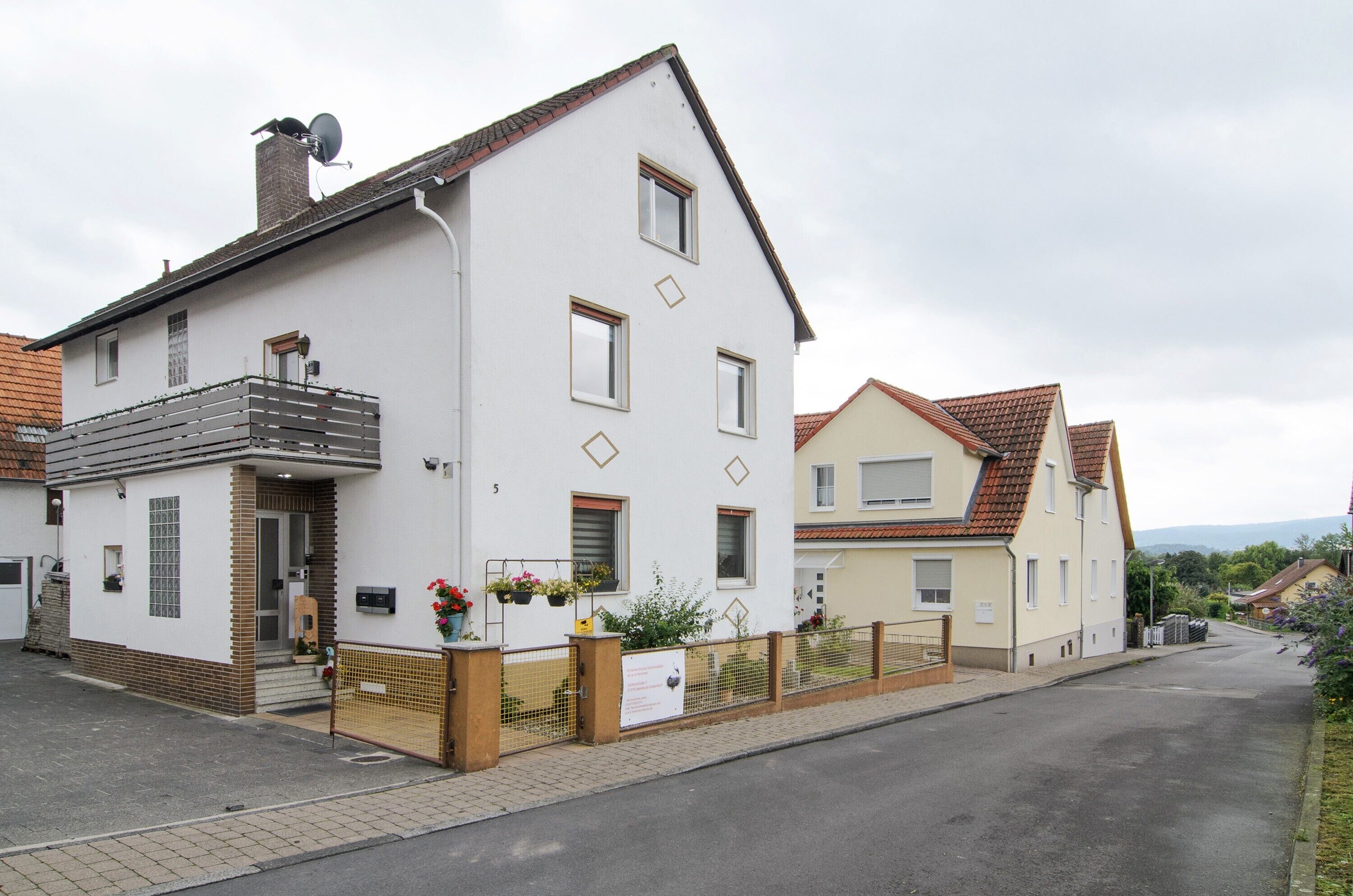 A two-story residential building with a balcony, a small porch, and a fenced front yard with potted plants.