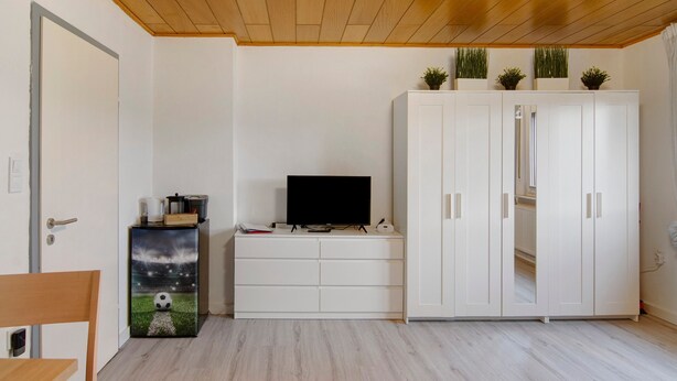 Una habitación con un armario blanco, un televisor sobre un mueble, una mesita y una puerta.
