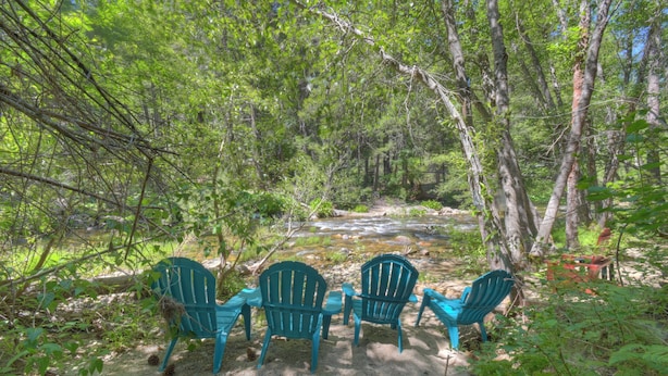 En vacker, naturlig miljö med en flod, omgiven av täta träd och tre Adirondack-stolar som är placerade med utsikt mot floden.