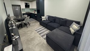 A modern living room with a grey sofa, a dining table with chairs, a kitchen area in the background, and a large flat-screen TV on a stand. - Modern Comfort in Oba: New 1+1 Apartment in Sol Garden (Alanya)