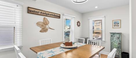 Un espace repas comprenant une table en bois, des chaises blanches, un élément décoratif mural et un luminaire au plafond.