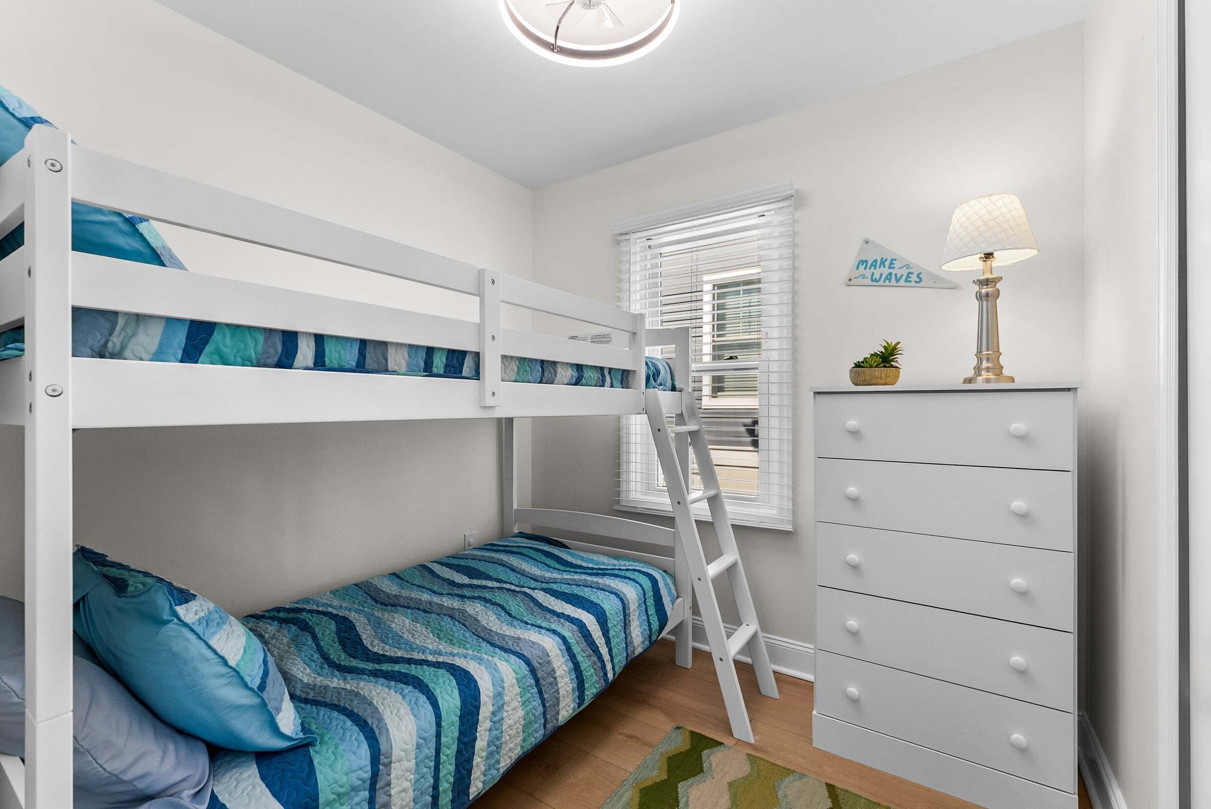 Ein Stockbett mit einer Leiter, eine Kommode mit Schubladen, eine Lampe und ein Fenster mit Jalousien.