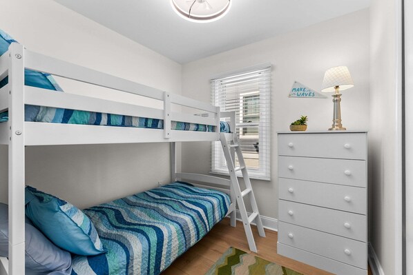 A bunk bed with a ladder, a dresser with drawers, a lamp, and a window with blinds.