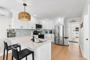 A modern kitchen with white cabinets, a central island with bar stools, a stainless steel refrigerator, and a woven pendant light.