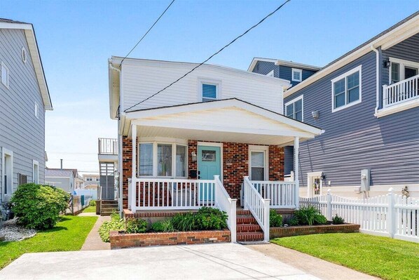 A two-story residential building with a front porch, brick foundation, and a white fence.