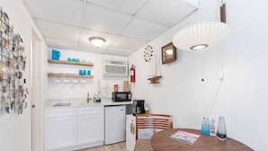 A compact kitchen with white cabinets, a microwave, a small fridge, and a dining table with chairs. - Top-Floor Studio with a Heated Pool & Parking (Wildwood)