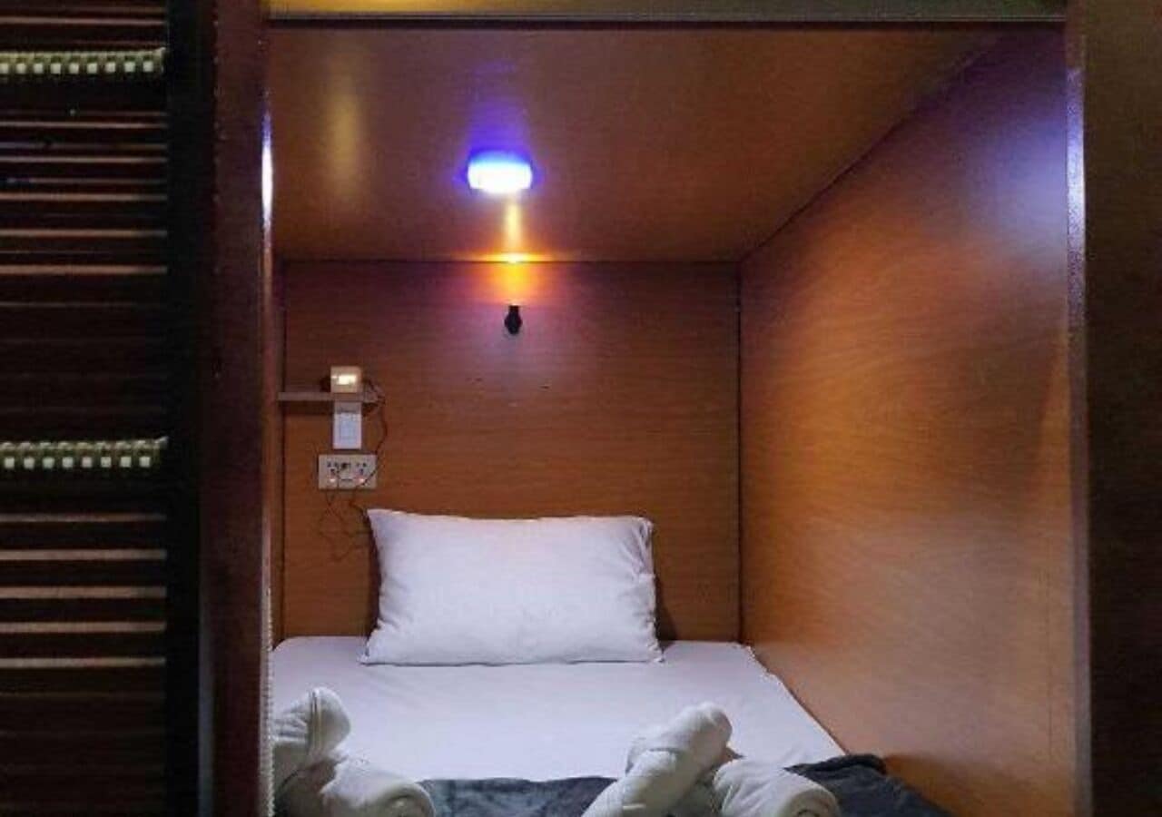 Una habitación pequeña, con poca luz, que tiene una cama individual, una mesita de noche y una lámpara fijada a la pared.