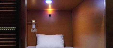 A small, dimly lit room with a single bed, a nightstand, and a wall-mounted light fixture.