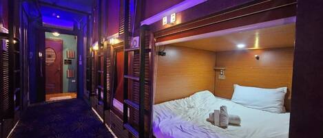 A narrow hallway with a patterned carpet leading to a room with a bed, wooden headboard, and a small window.