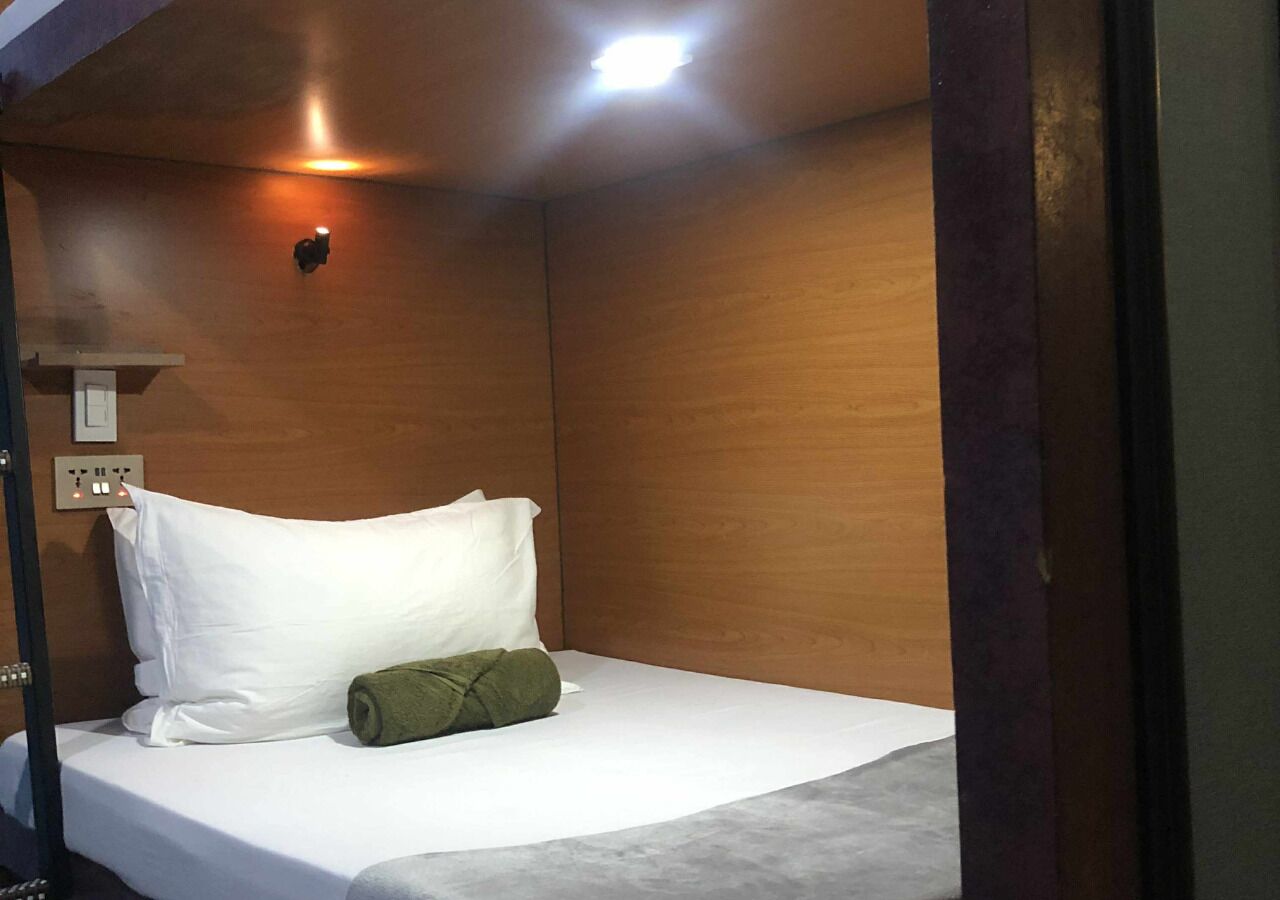A single bed with white bedding and a green towel, a wall-mounted light, and a power outlet.