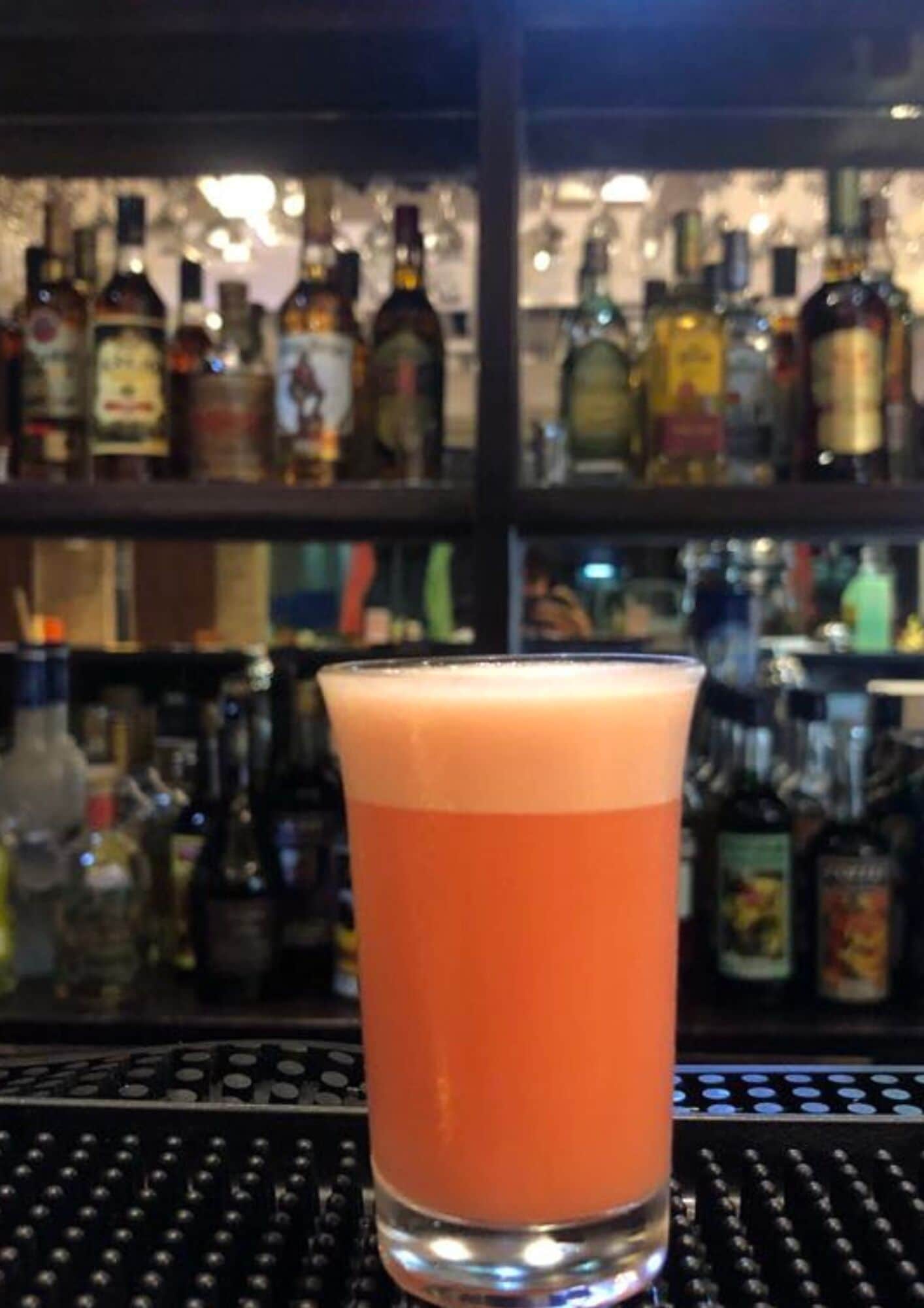 A tall glass of orange-colored beverage on a bar counter with a blurred background of various bottles.
