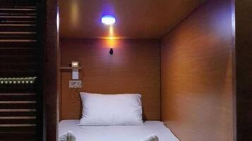 A small, enclosed room with a single bed, a pillow, and two rolled-up towels. There is a wall-mounted light fixture above the bed.
