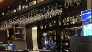 Baarialue, jossa on vastaanottotiski, hyllyt täynnä alkoholijuomapulloja ja katosta roikkuvia viinilaseja.