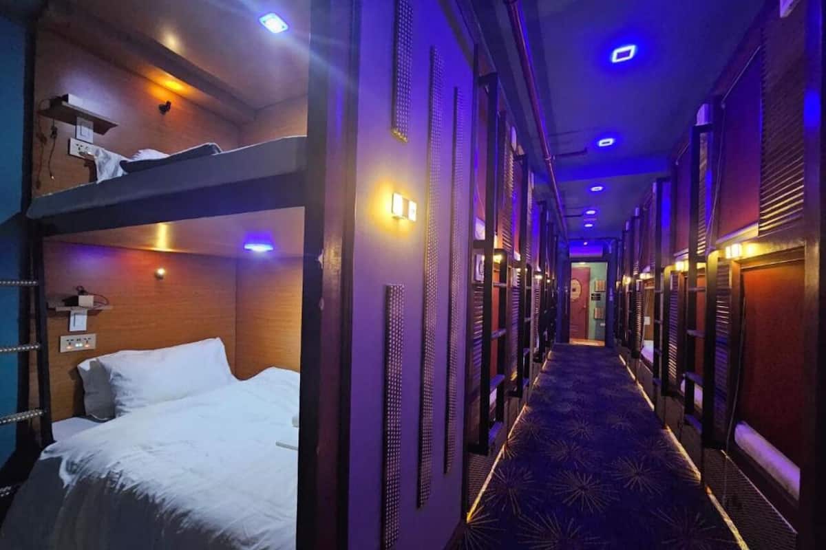 A hotel corridor with bunk beds on the left, a patterned carpet, and a long hallway with doors on the right.