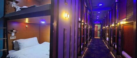A hotel corridor with bunk beds on the left, a patterned carpet, and a long hallway with doors on the right.