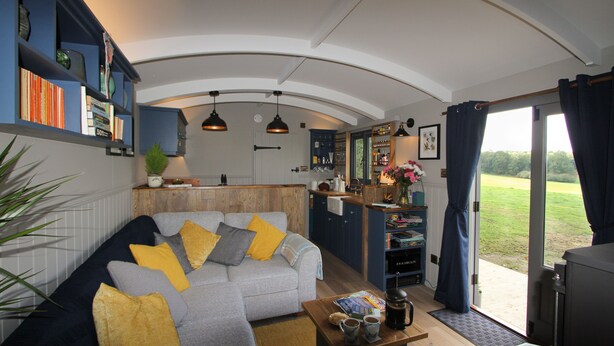 A modern living room with a grey sofa, yellow cushions, and a coffee table. There is a kitchen area with wooden cabinets and a dining table. The room has a large sliding glass door leading to a garden. - Top Farm Shepherds Lodge, nestled in a peaceful farm in Marsham, North Norfolk (Marsham)