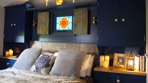 A bedroom with a large bed, wooden headboard, and a stained glass window above the bed. There are bedside tables with candles and a small shelf with decorative items. - Top Farm Shepherds Lodge, nestled in a peaceful farm in Marsham, North Norfolk (Marsham)