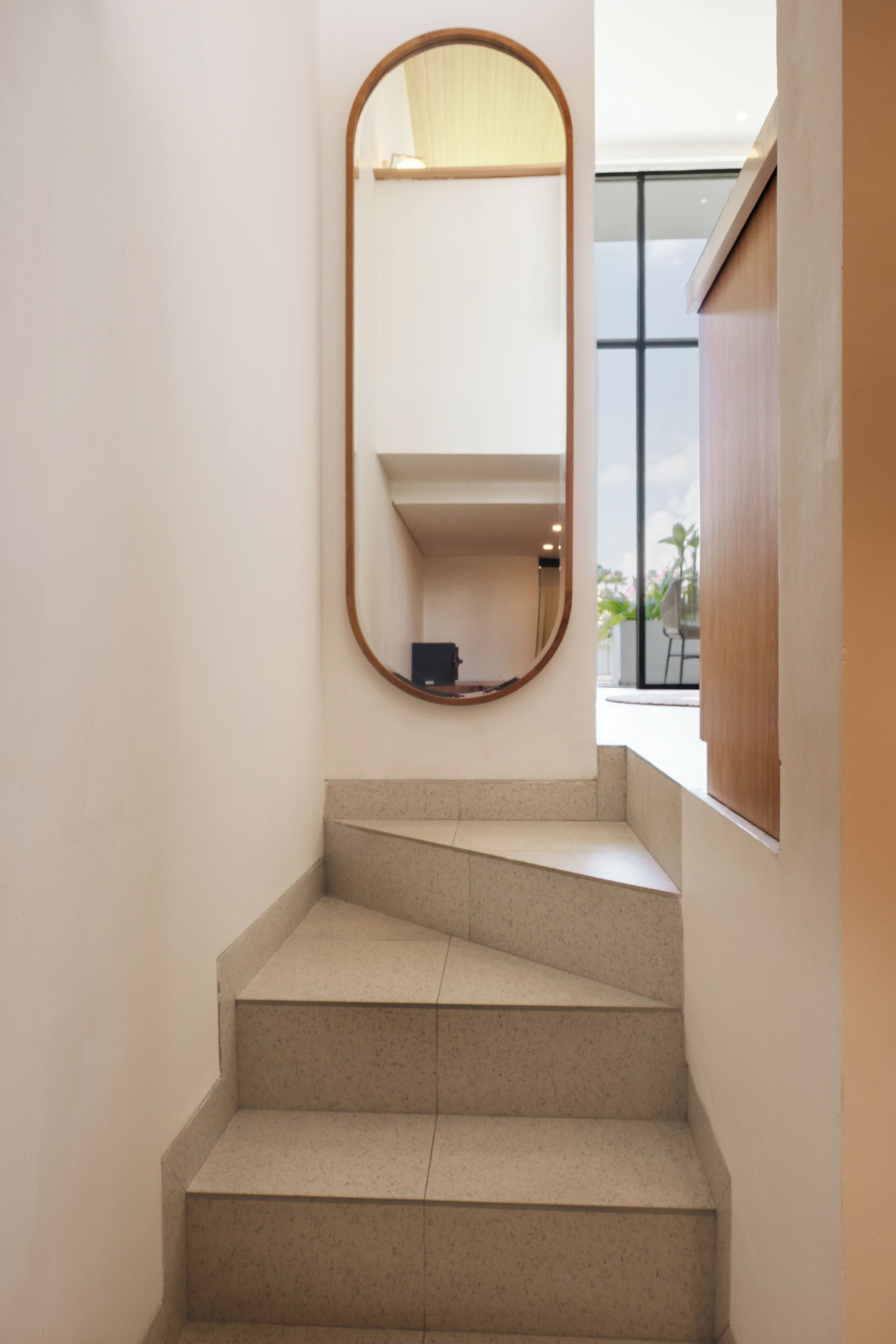A staircase with a wooden handrail, a mirror on the wall, and a window with a view of greenery.