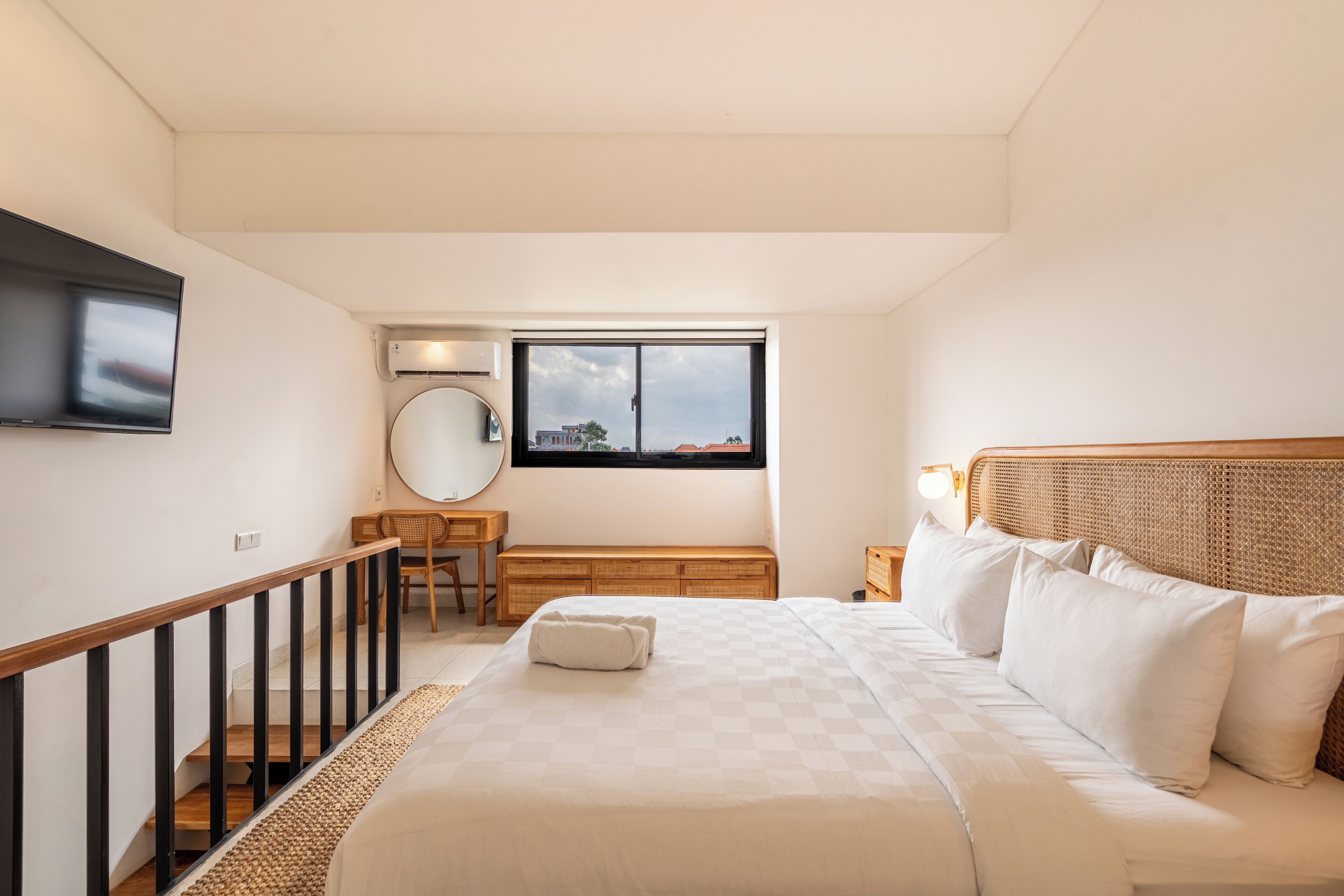 A bedroom with a bed, a desk, a chair, a round mirror, and a window with a view of buildings.