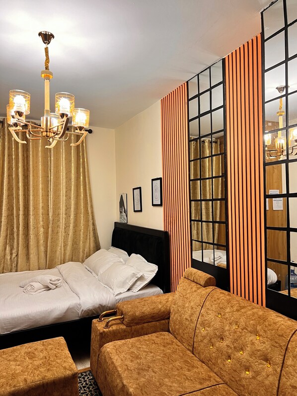 Une chambre d’hôtel avec un lit, un canapé, un lustre, des rideaux et une fenêtre avec un store à rayures.