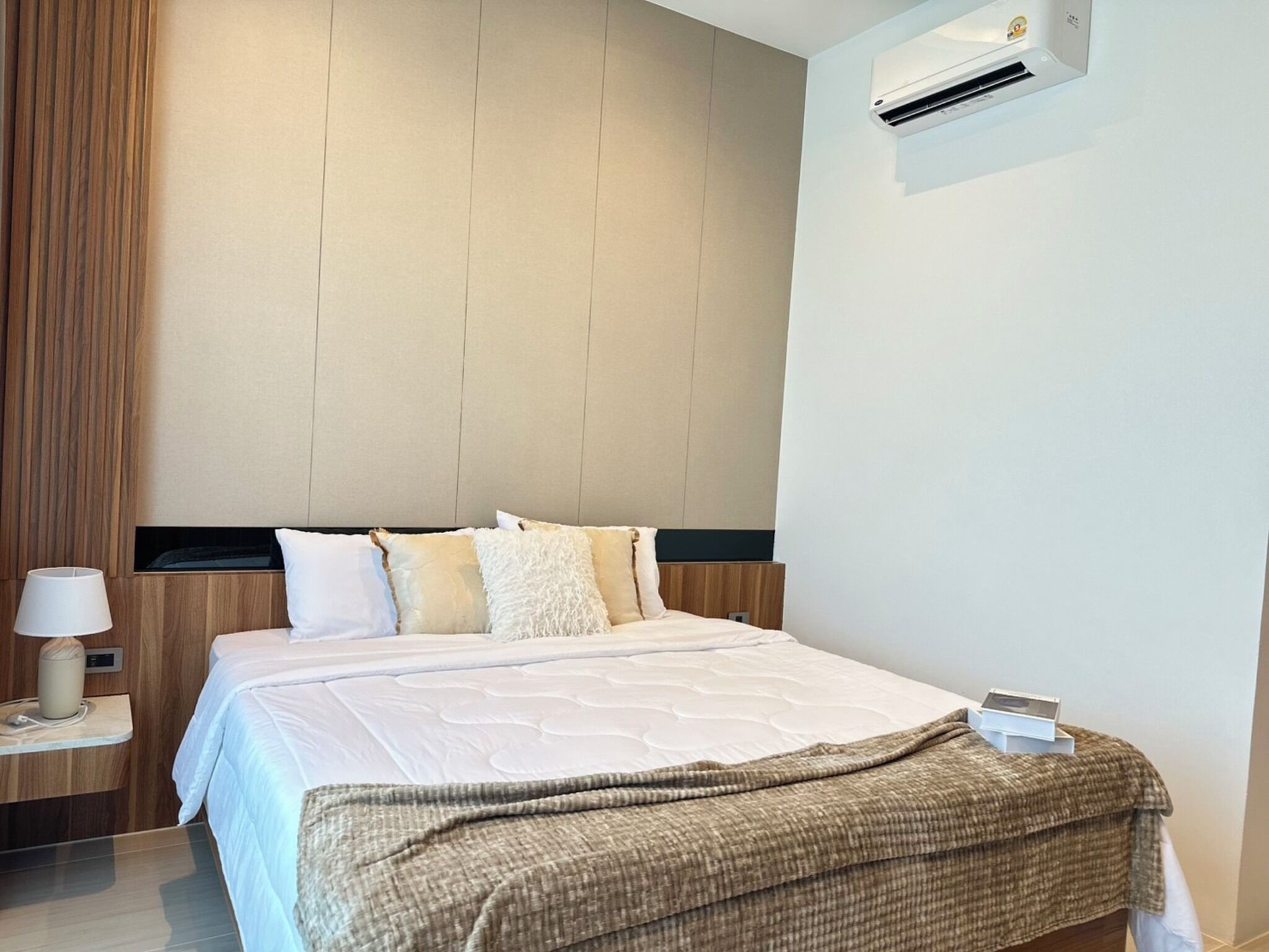 A modern hotel room with a large bed, a bedside table with a lamp, and a wall-mounted air conditioner.